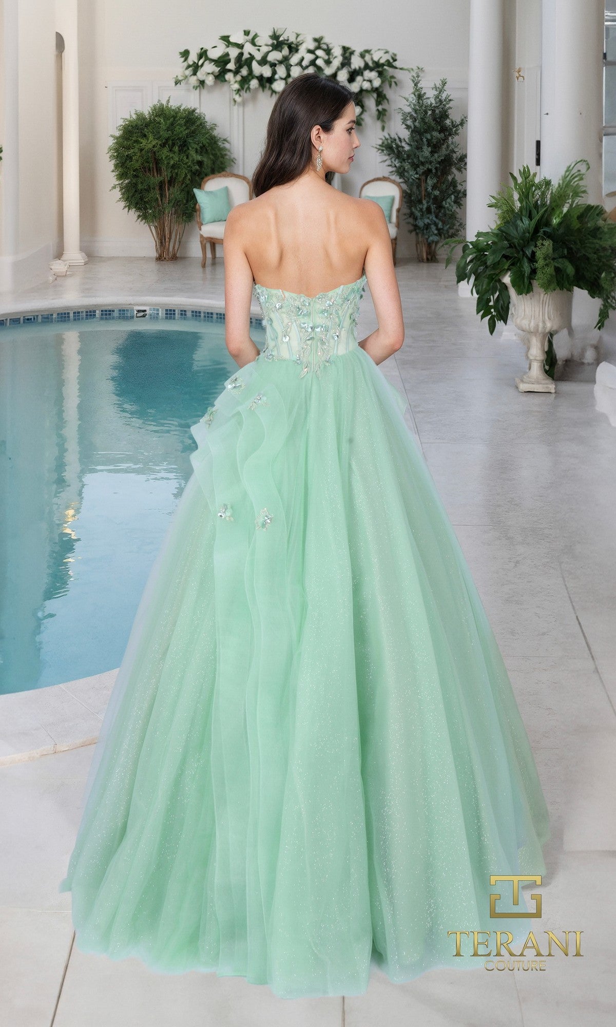 A woman in the Terani 261P6197 long formal dress stands by an indoor pool, facing away. The mint green strapless gown has a full tulle skirt, floral accents, and sequin-embellished lace. The Terani Couture logo is visible.